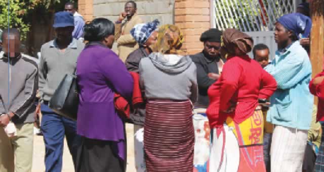 Curious neighbours gather outside the Pumula South house in Bulawayo where the murder took place while a police officer leads the teenage suspect away yesterday